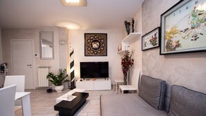 Een moderne woonkamer met een flatscreen televisie, een bank, een salontafel en decoratieve elementen, waaronder een spiegel, ingelijste kunstwerken en een potplant.