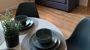 Una sala moderna con un sofá gris, una mesita auxiliar con una lámpara, y un comedor con mesa dispuesta para dos.