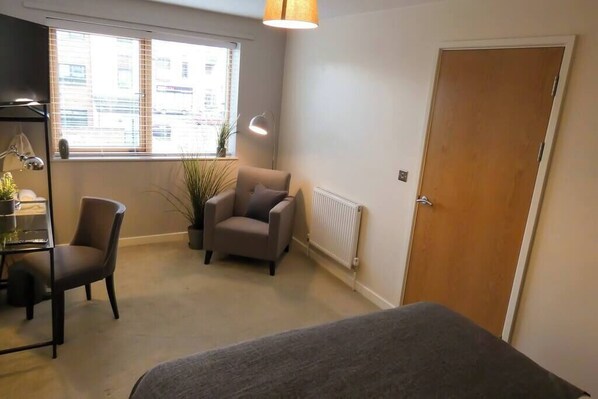 A room with a bed, a desk with a chair, a potted plant, a window with blinds, and a radiator.