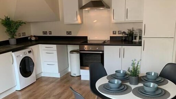 Una cocina moderna con armarios blancos, horno empotrado, lavadora y comedor con mesa redonda y sillas.