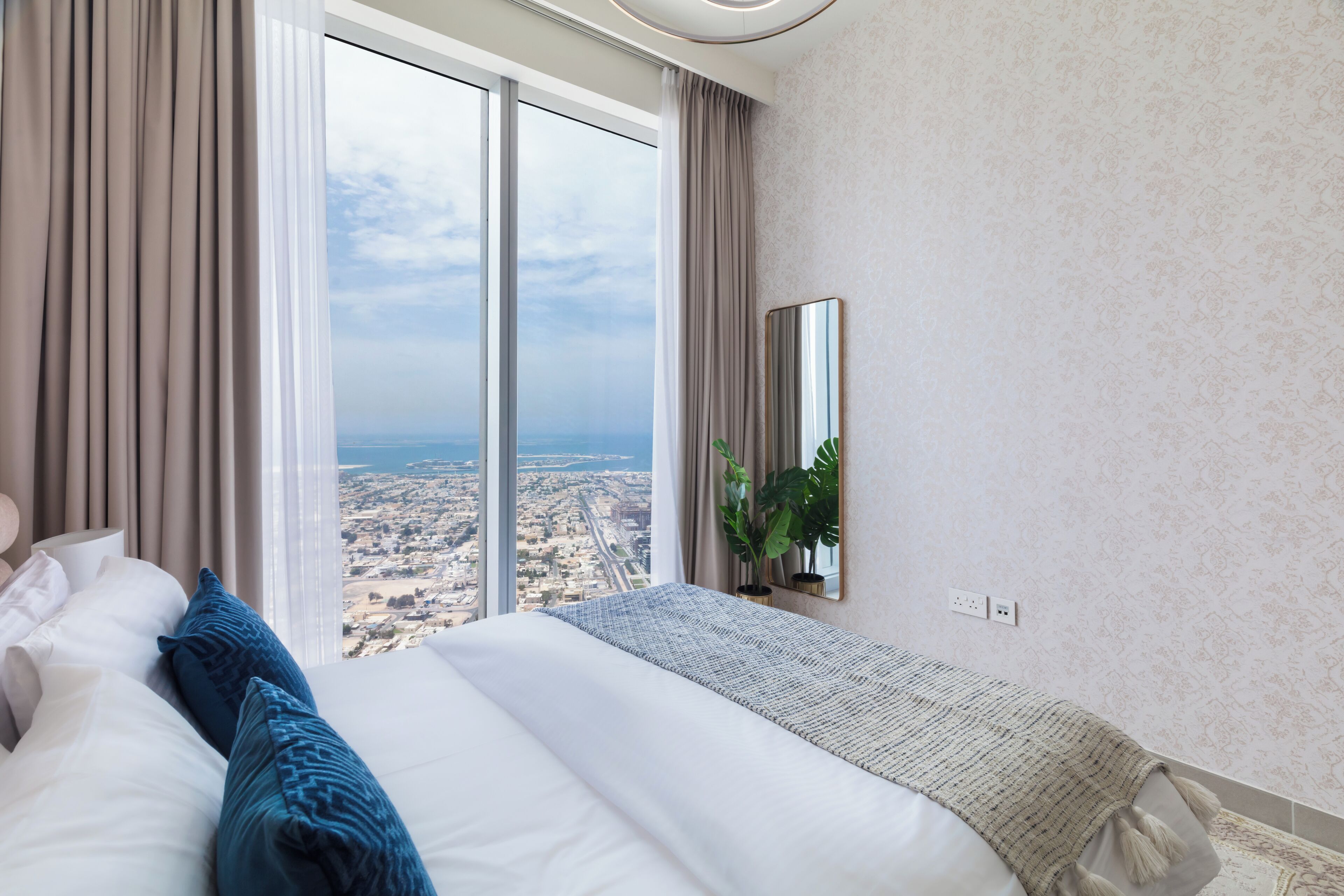 Une chambre d’hôtel moderne avec un grand lit, une vue sur le paysage urbain grâce à de grandes fenêtres du sol au plafond, un miroir au mur et une plante sur le rebord de la fenêtre.