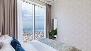 Une chambre d’hôtel moderne avec un grand lit, une vue sur le paysage urbain grâce à de grandes fenêtres du sol au plafond, un miroir au mur et une plante sur le rebord de la fenêtre.