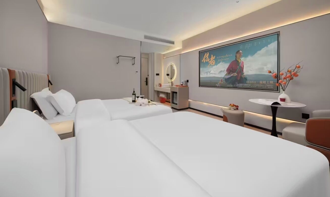 Hotellihuoneessa on kaksi sänkyä, peilillä varustettu meikkipöytä, pieni pöytä maljakolla ja juliste seinällä.