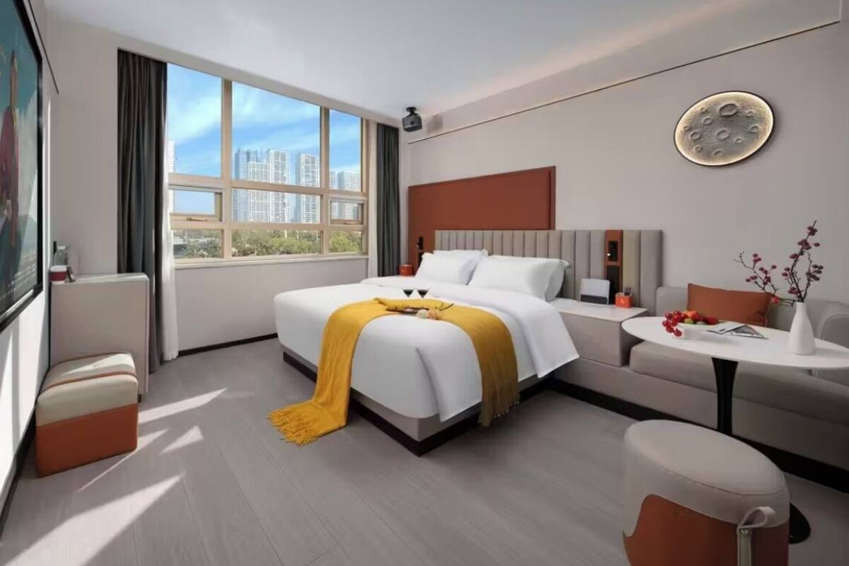 A modern hotel room with a large bed, a sofa, a small table, and a view of the cityscape through large windows.