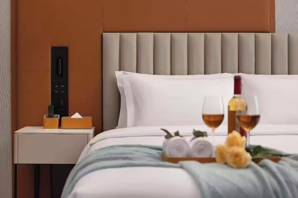 A hotel room with a bed, a nightstand with a phone and a tissue box, and a tray with a bottle, glasses, and rolled towels.