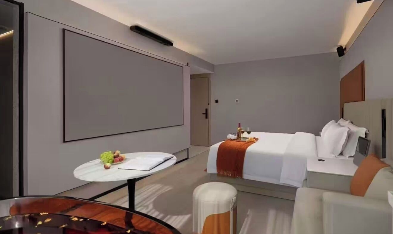 Une chambre d’hôtel moderne avec un grand lit, une petite table ronde sur laquelle sont disposés des fruits, un canapé et un écran de télévision.