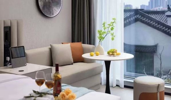 A modern living room with a sofa, a small round table with a vase and fruit, a laptop on a side table, and a view of traditional architecture through a window. - Hefei Yunmao Art Hotel (Hefei)