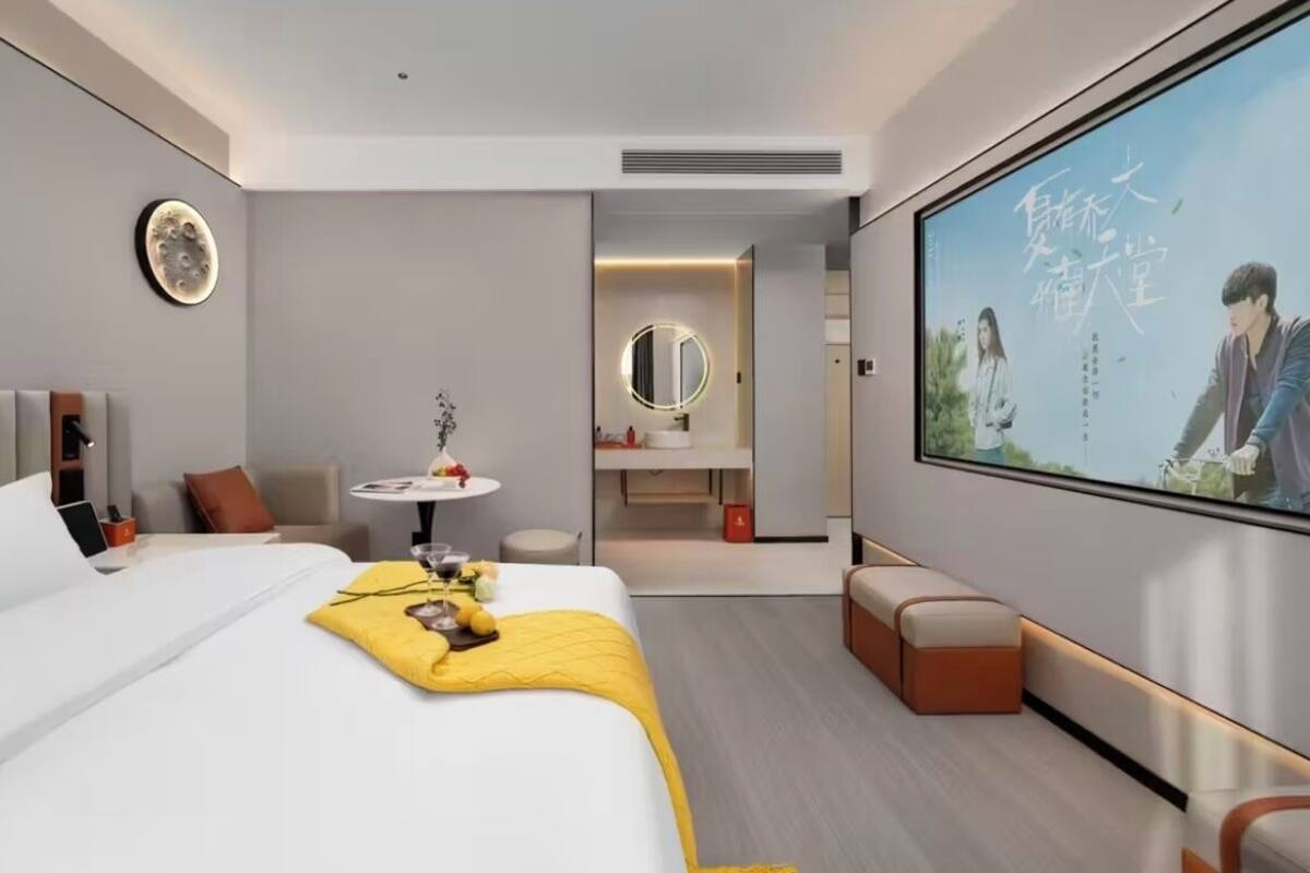 A modern hotel room with a large bed, a small round table with a vase, a TV screen, and a seating area with cushions.