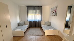 Une petite pièce bien éclairée, avec deux lits simples, une grande fenêtre avec des rideaux, un plancher en bois et un tableau accroché au mur.