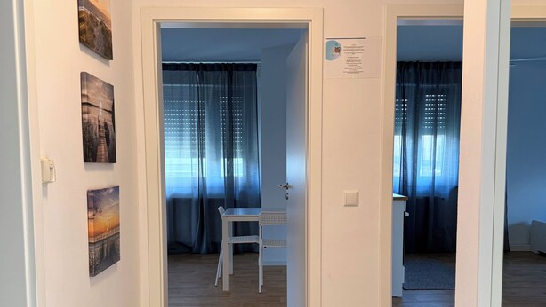 Un couloir doté d’un plancher en bois, de murs blancs et d’une porte donnant sur une pièce où se trouvent une table et une chaise.