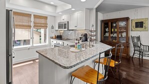 A modern kitchen with white cabinets, granite countertops, and stainless steel appliances. There is a breakfast bar with bar stools and a wooden cabinet with glass doors. - Great Beach Condo in Quiet Section of Wild Dunes Resort! *301 Yacht Harbor* (Isle Of Palms)
