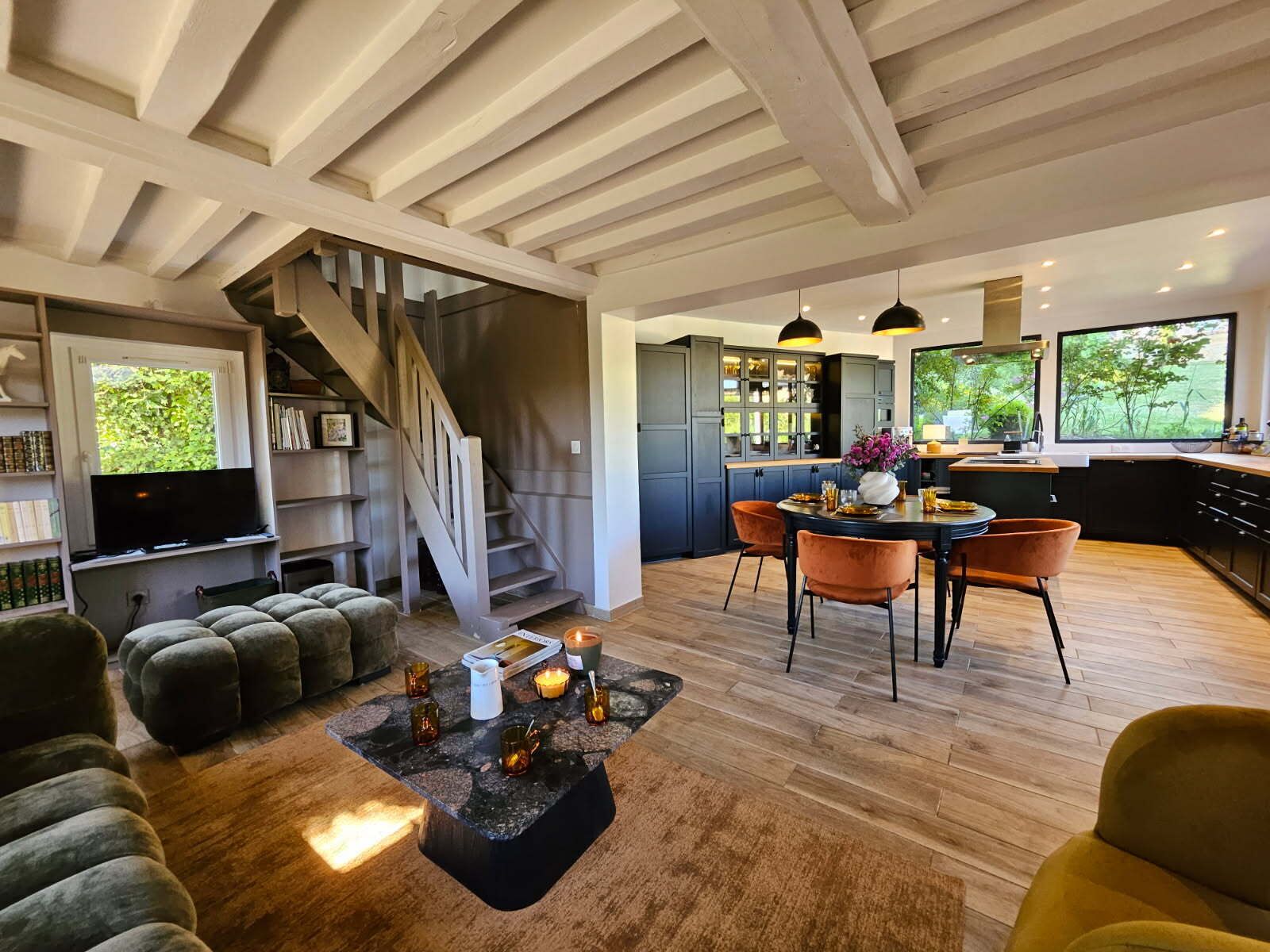 Een moderne woonkamer met een open haard, een eethoek met oranje stoelen en een keuken met donkere kastjes en een groot raam.