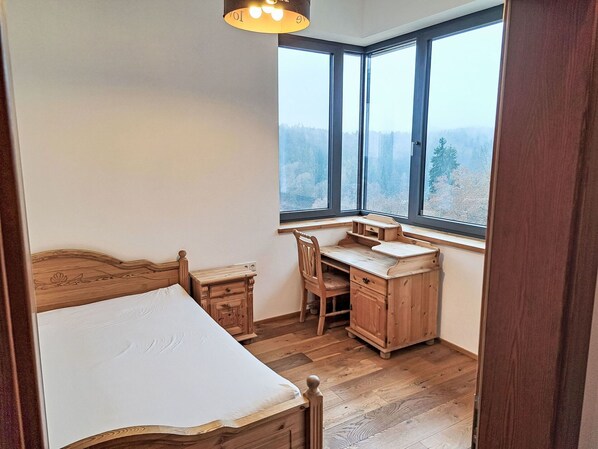 Ein Schlafzimmer mit einem hölzernen Bett, einem Schreibtisch und einem Stuhl, einem Fenster mit Blick auf Bäume und einer Deckenleuchte.