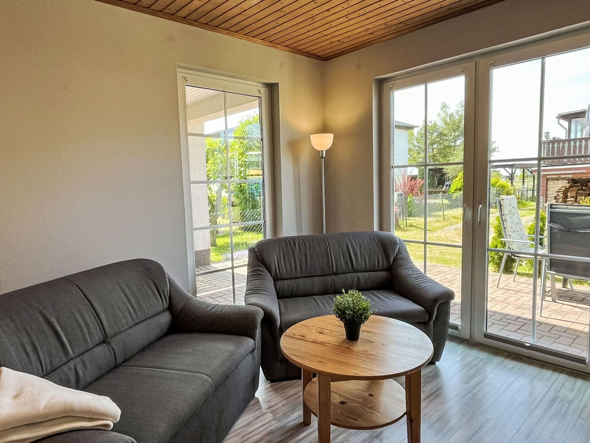 A modern living room with a grey sofa, a round wooden coffee table with a plant, and large sliding glass doors leading to an outdoor patio area.