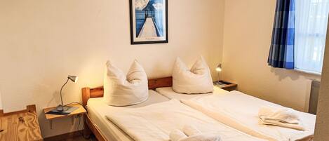 Cama doble con ropa de cama blanca, dos almohadas, mesita de noche con lámpara, cuadro enmarcado en la pared y ventana con cortinas.