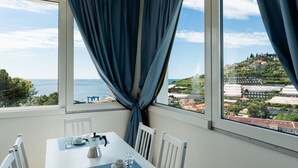A dining area with a table set for two, a coffee maker, and a view of the sea and a hill with buildings. - Cosy apartment for 6 guests with WIFI, A/C, TV, balcony and pets allowed (Sanremo)