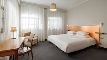 A hotel room with a bed, a desk, a chair, and a lamp.