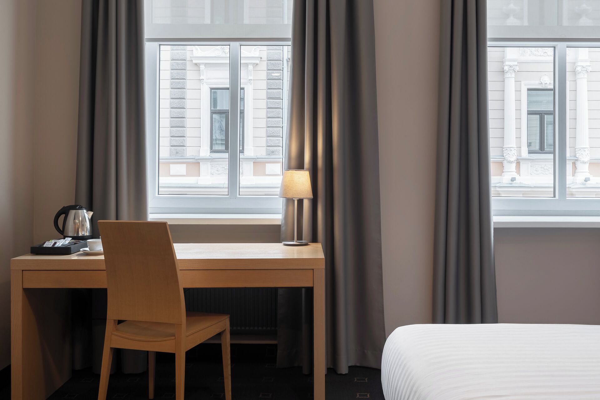 Une chambre d’hôtel avec un bureau en bois, une chaise, une lampe, un lit et une fenêtre avec des rideaux.
