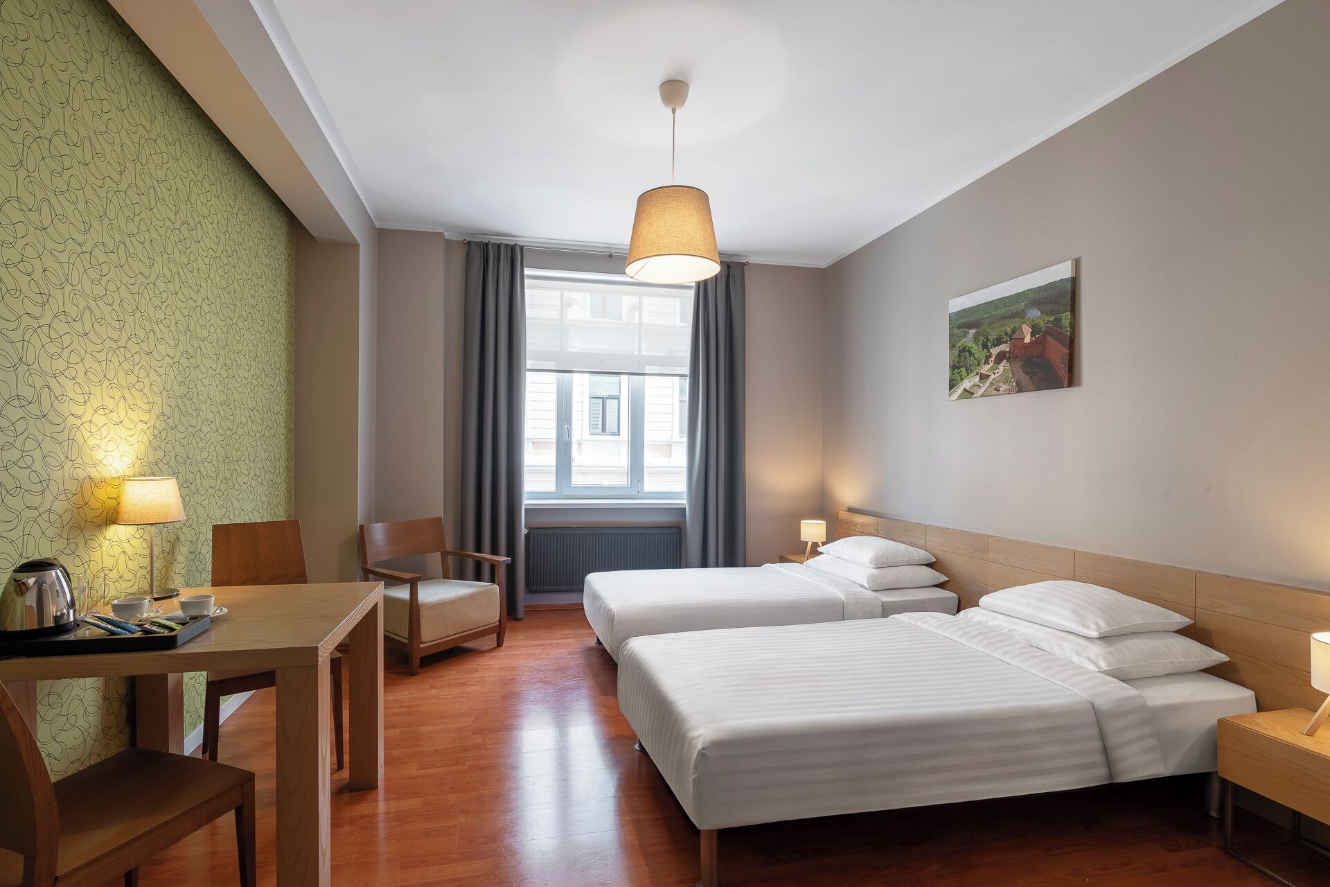 A hotel room with two beds, a desk with a kettle, a chair, and a view of a building through the window.