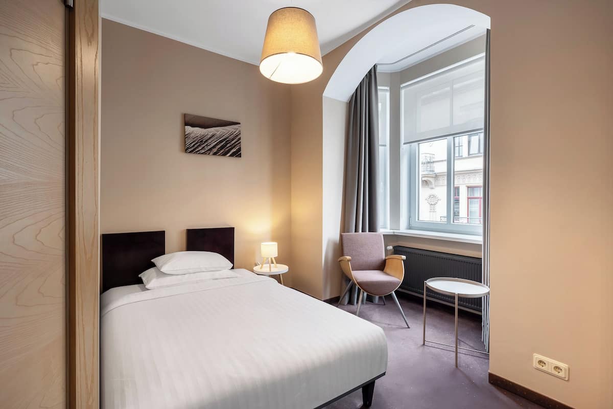 Habitación de hotel con cama, una mesita, una silla, lámpara, ventana y un cuadro en la pared.