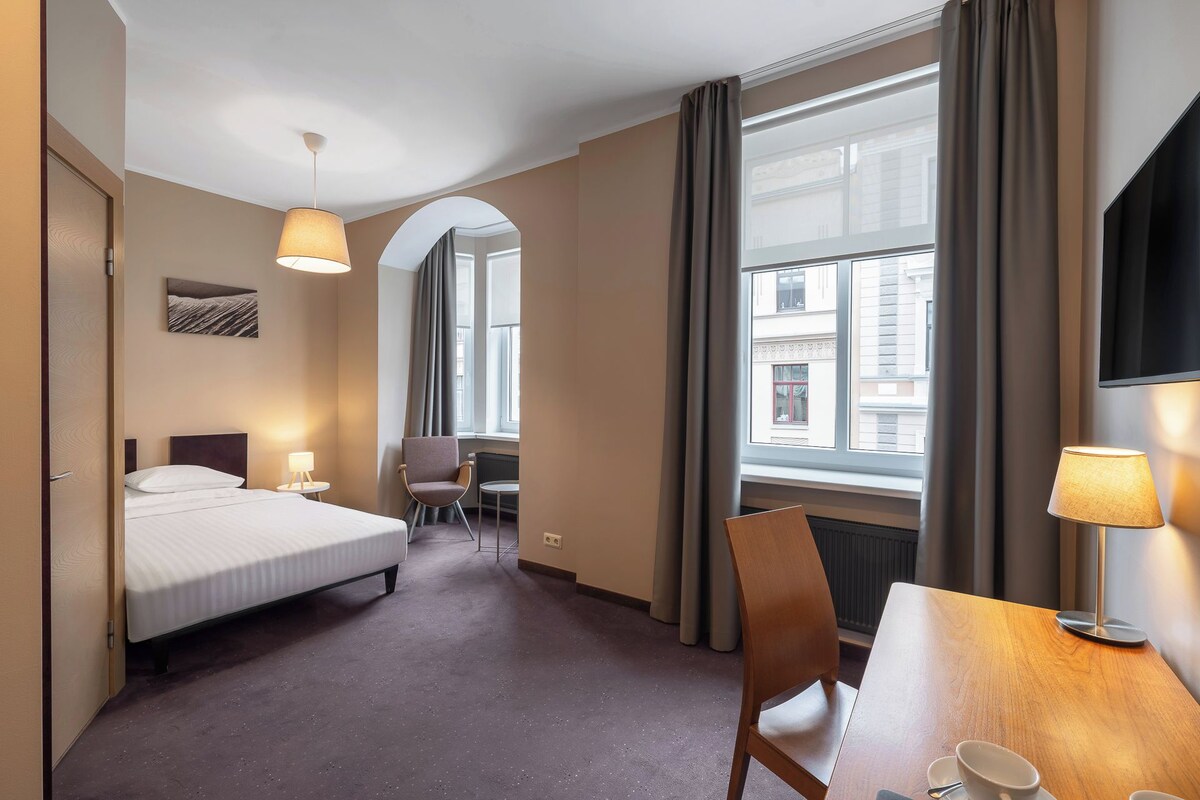 Una habitación de hotel con cama, una silla, una mesa con una taza y una ventana con cortinas.