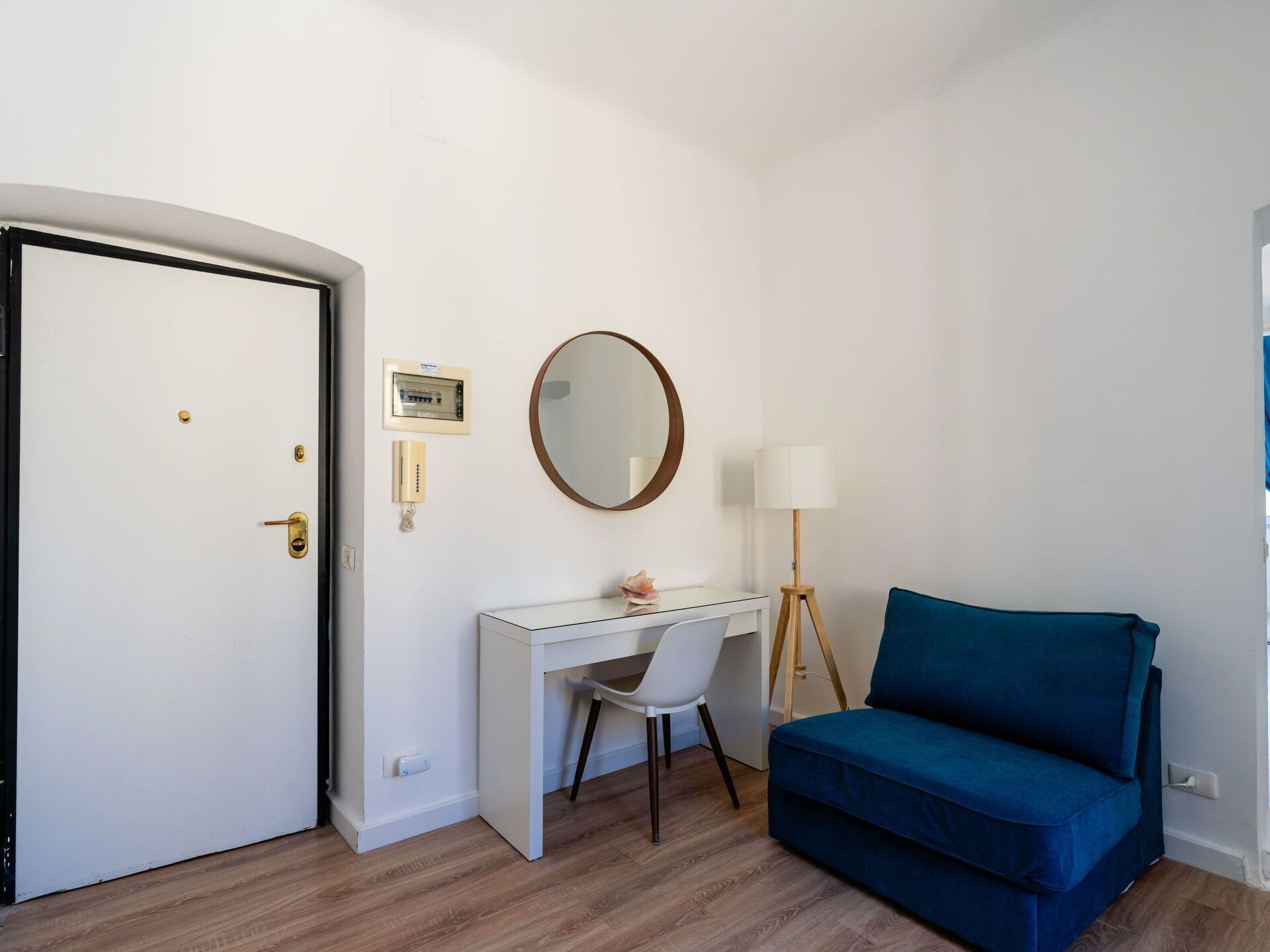 A room with a white door, a desk with a chair, a blue sofa, a round mirror, a lamp, and a wall-mounted phone.
