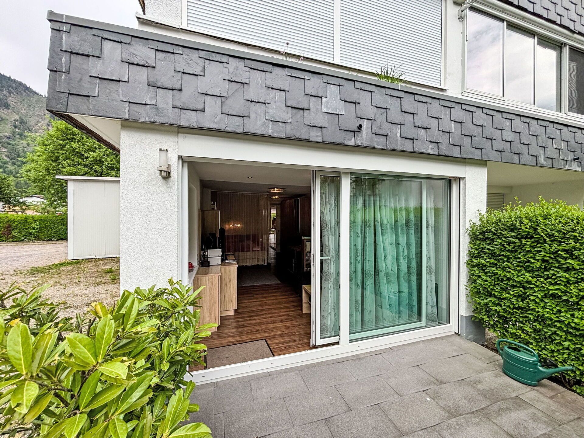 A modern house with a sliding glass door entrance, a wooden staircase leading to an upper floor, and a neatly trimmed hedge.