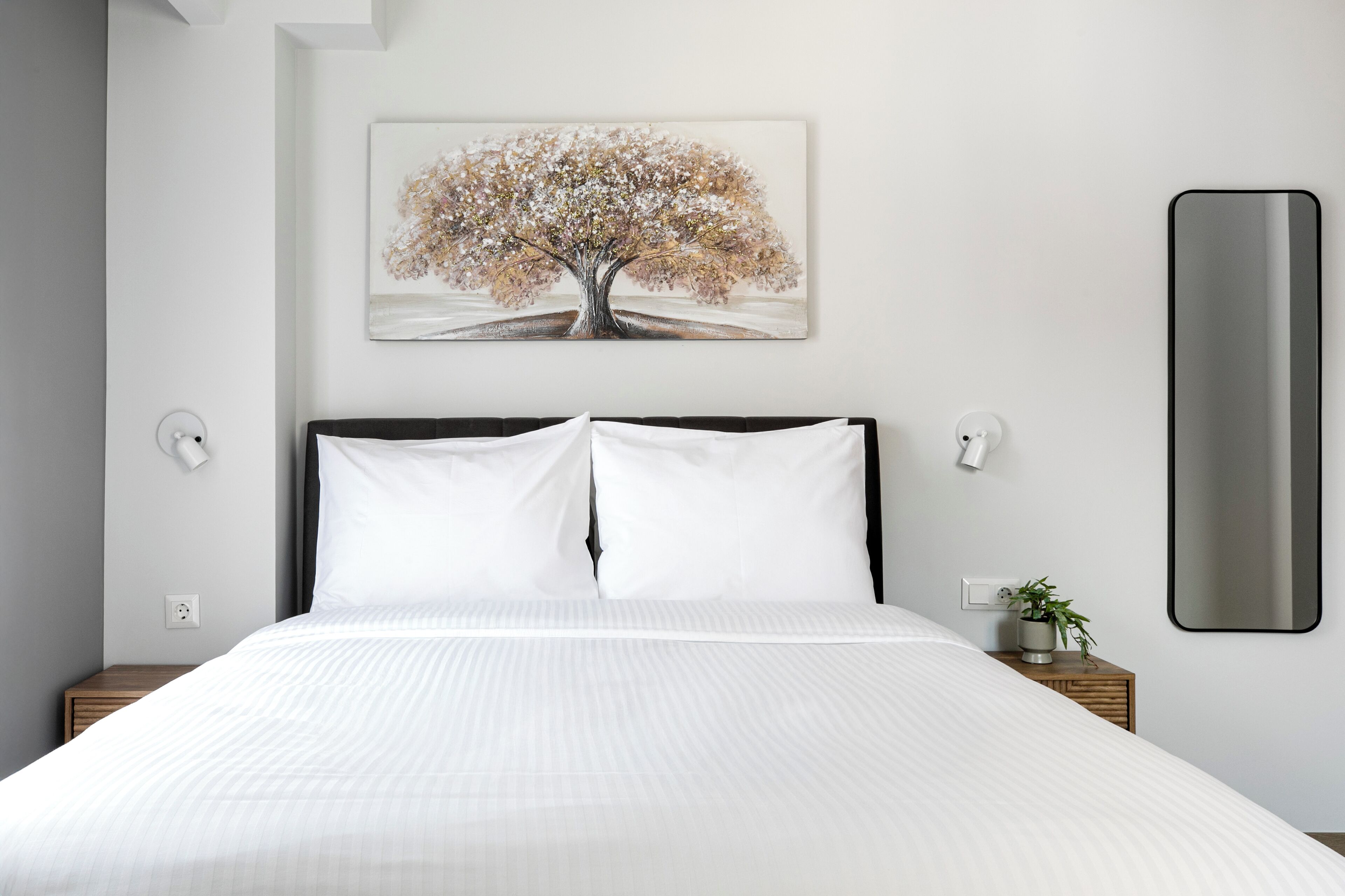Una cama bien hecha con sábanas blancas, un cuadro grande de un árbol en la pared y una mesita de noche con una planta.