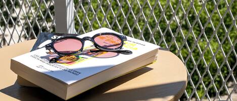 A round table with a book and sunglasses on it, set against a metal fence with greenery in the background.