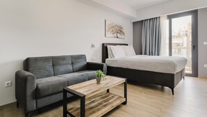 A modern bedroom with a grey sofa, a wooden coffee table with a plant, a bed with white bedding, and a city view through the window. - B48 Athens Dafni (Dafni-Ymittos)