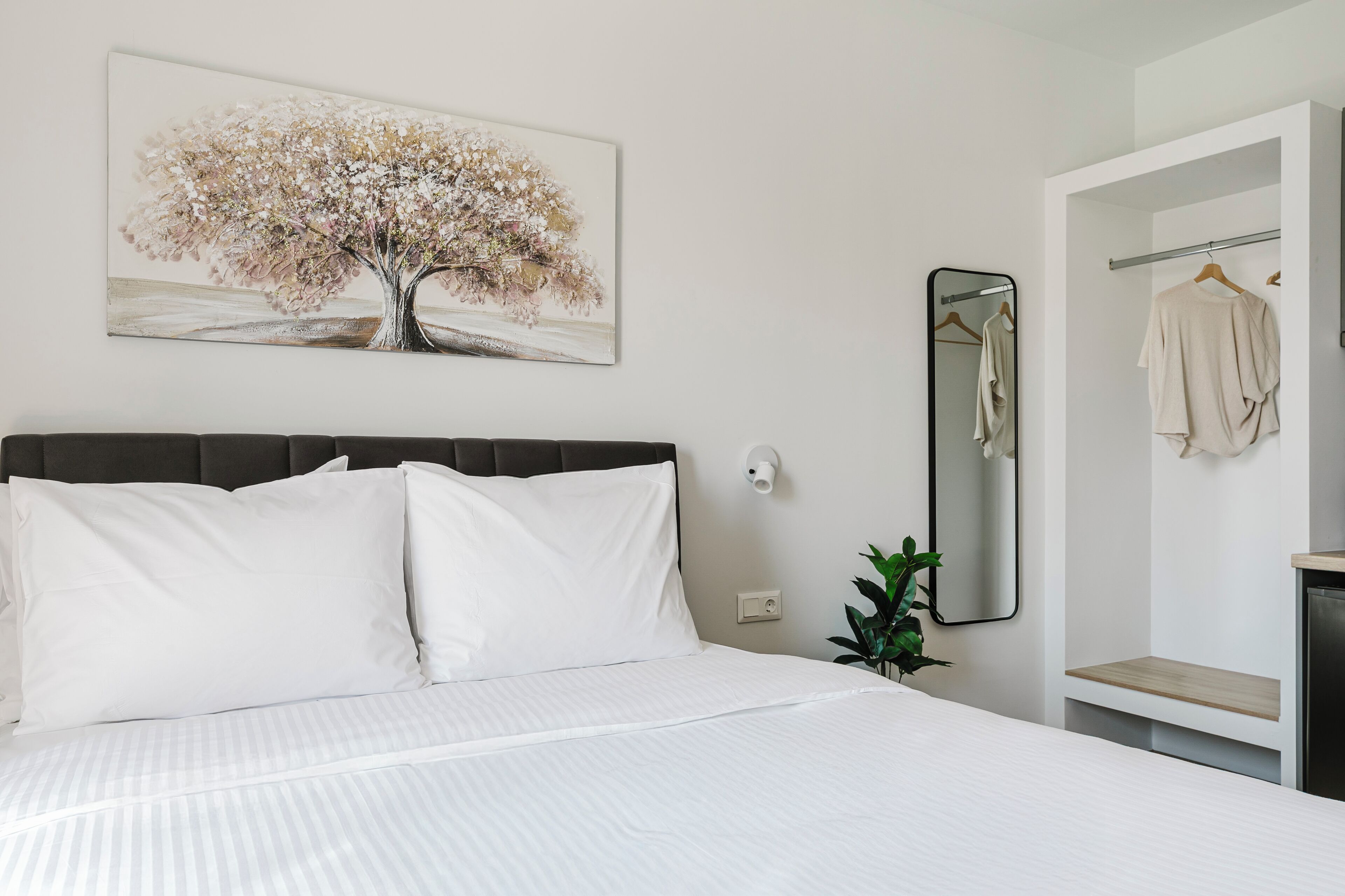 Une chambre à coucher avec un lit, du linge de lit blanc, un miroir fixé au mur, une robe suspendue et un tableau représentant un arbre.