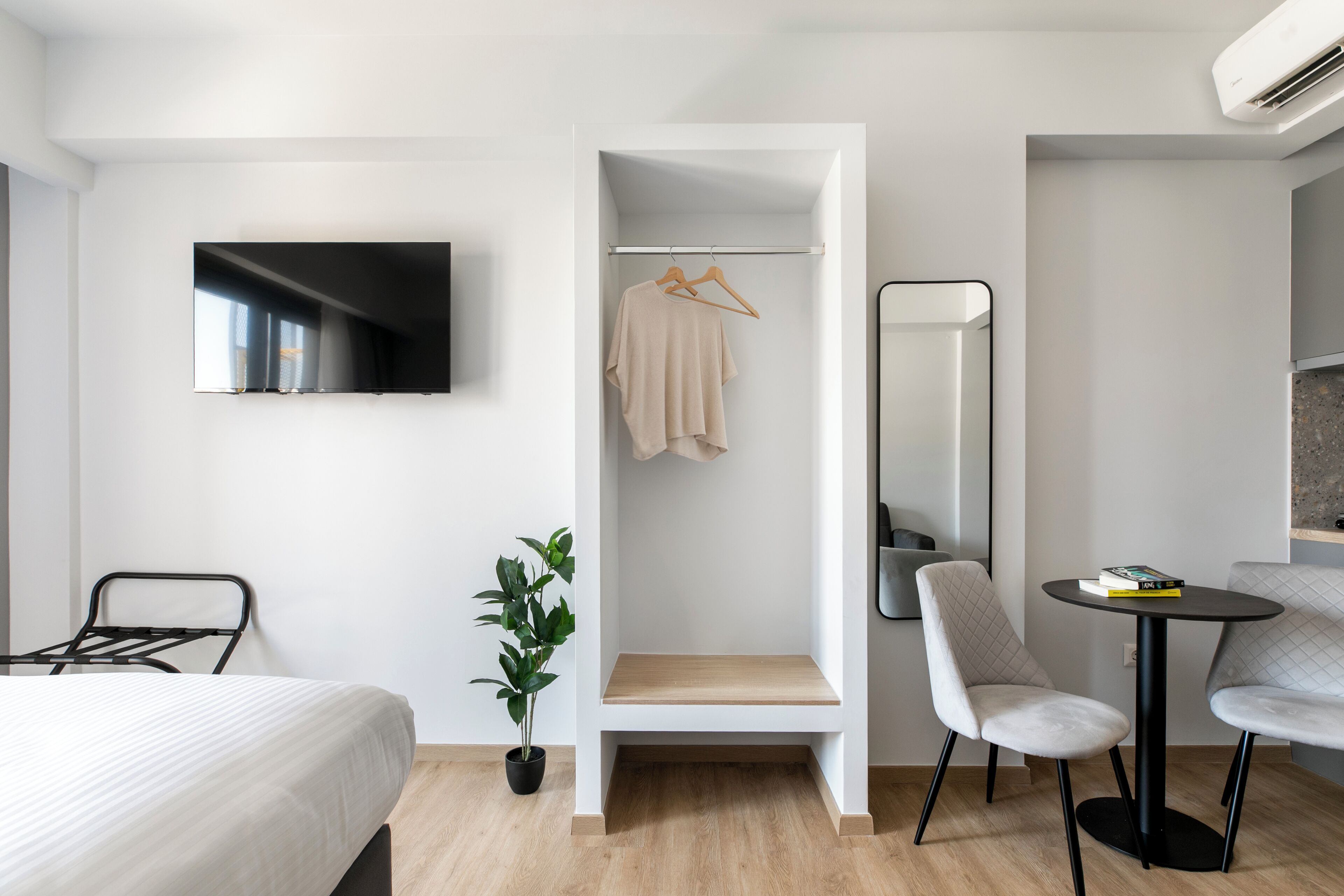 Habitación de hotel moderna con una cama, un área de descanso con mesa y sillas, un televisor montado en la pared, un armario con ropa colgada, una planta en maceta y un espejo.