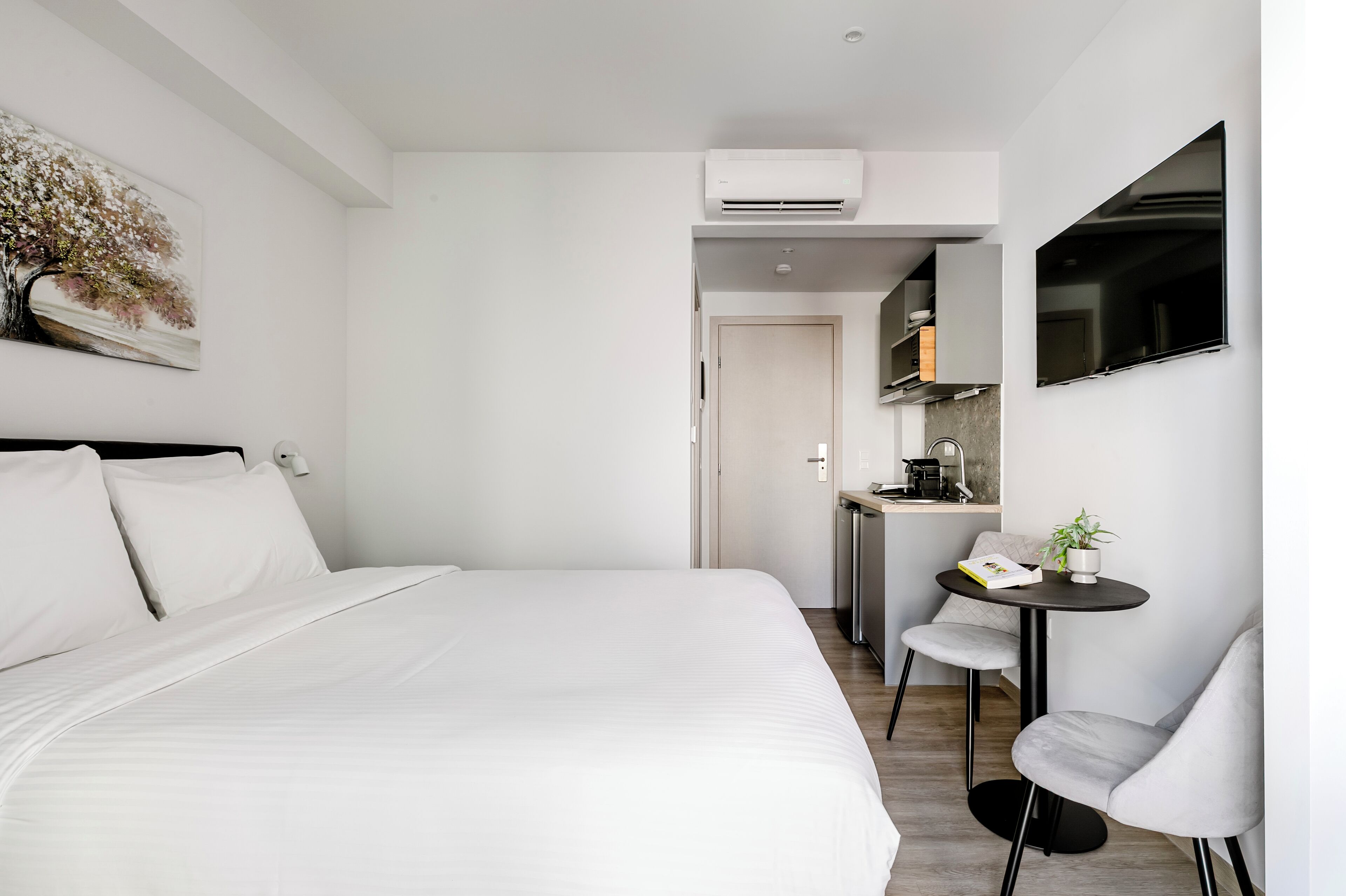 Ein modernes Hotelzimmer mit einem großen Bett, einer kleinen Küchenzeile, einem Essbereich und einem an der Wand befestigten Fernseher.