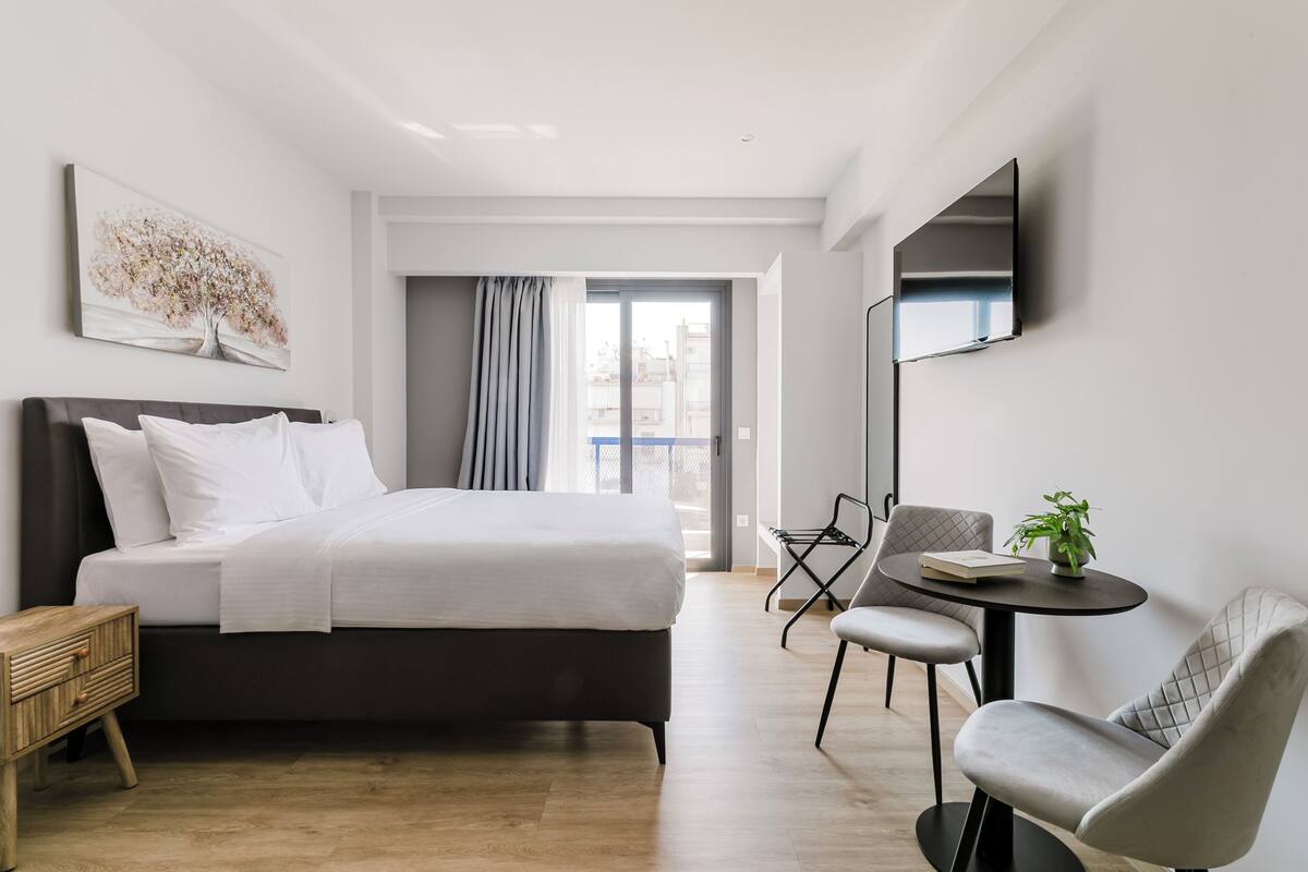 Ein modernes Hotelzimmer mit Bett, Schreibtisch und Stuhl. Es gibt ein Fenster mit Vorhängen, einen an der Wand befestigten Flachbildfernseher und eine Pflanze auf dem Schreibtisch.
