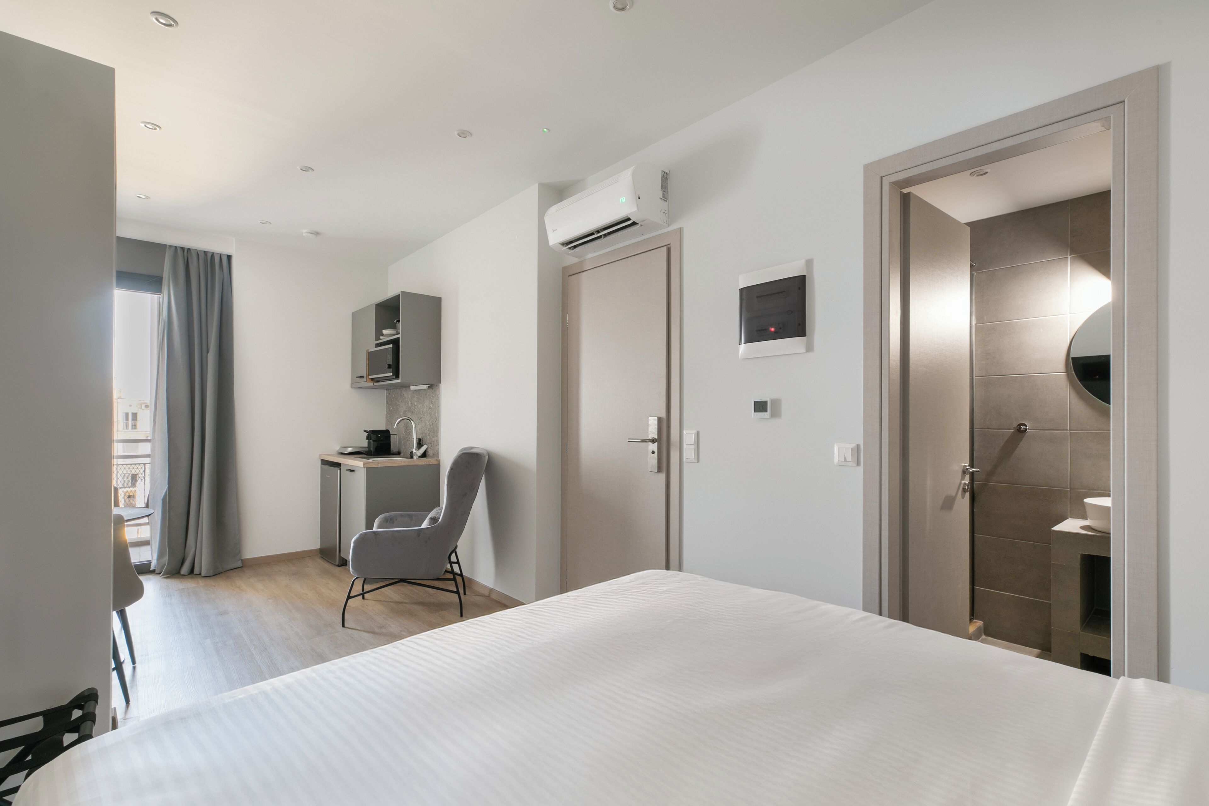 A modern hotel room with a large bed, a desk with a chair, an air conditioning unit, and a bathroom with a shower and toilet.