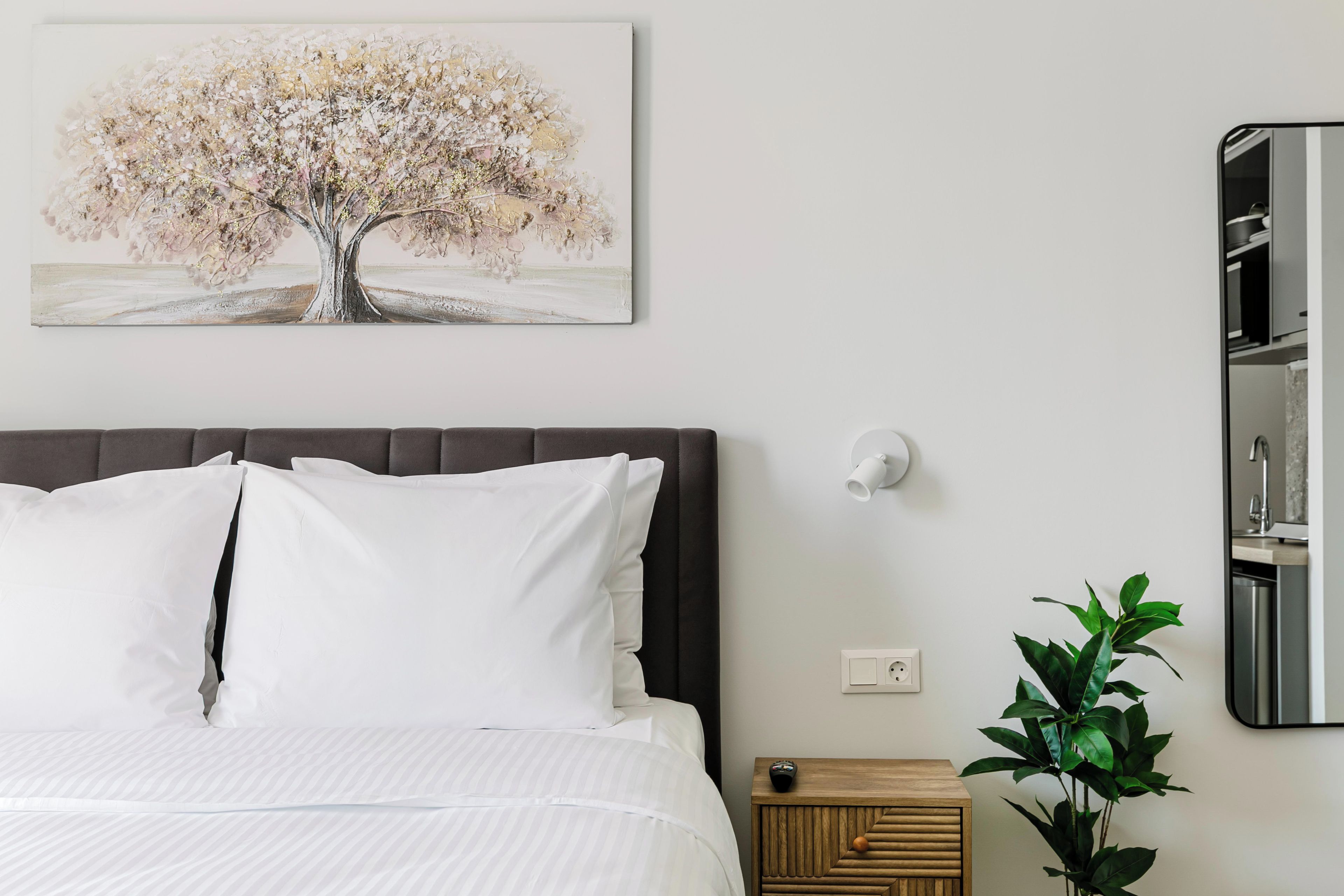 A bedroom with a bed, a bedside table, a wall-mounted light, a painting of a tree, and a potted plant.