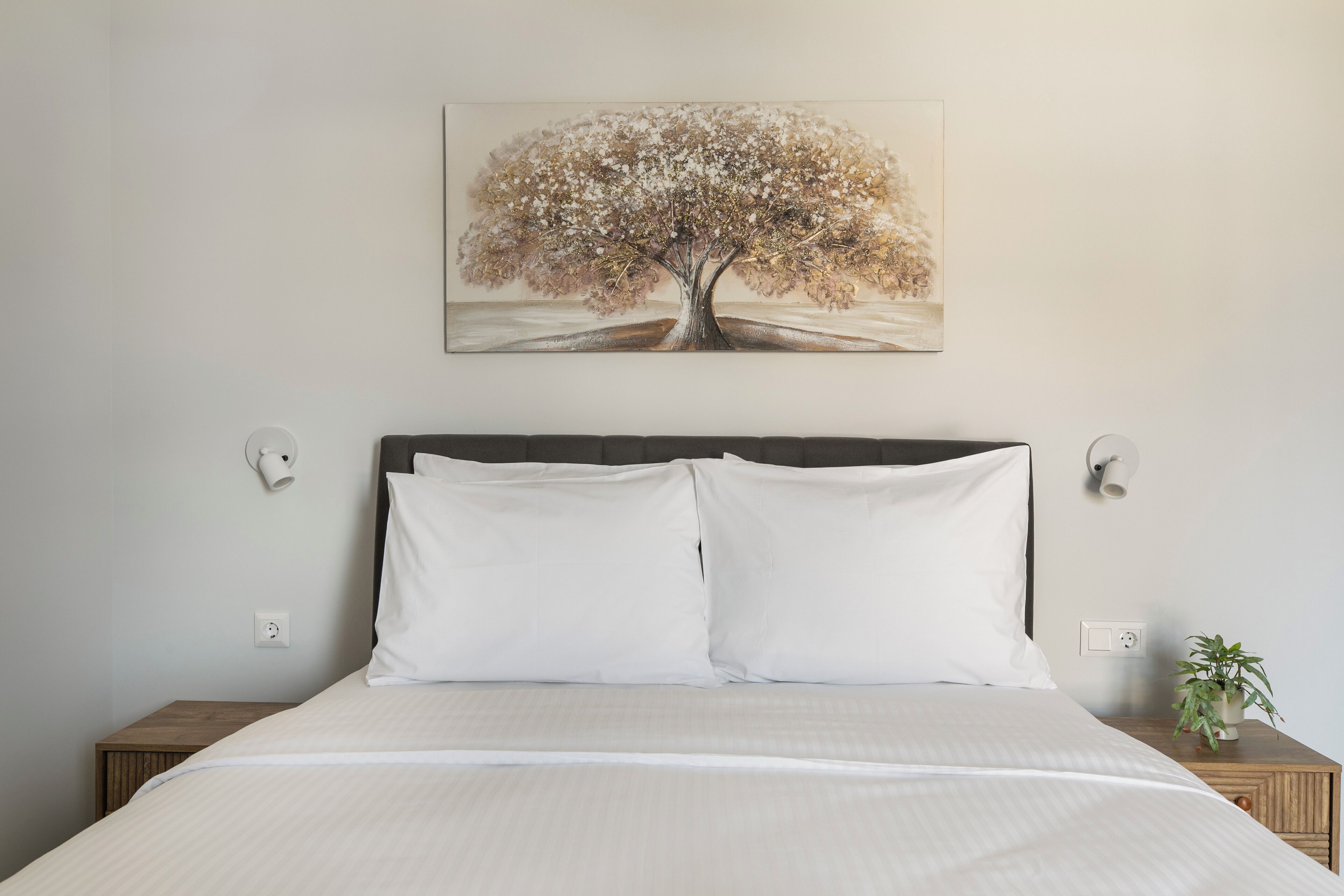 A neatly made bed with white linens, a dark headboard, and a wall-mounted painting of a tree.