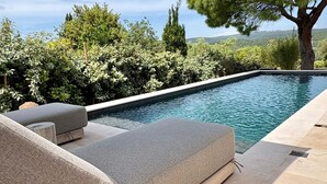 Et rektangulært pool med en klar blå vandoverflade, omgivet af en stendæk og en række grå loungesenge.