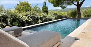 Una piscina rettangolare con superficie d'acqua cristallina, circondata da un solarium in pietra e una fila di poltroncine grigie imbottite.