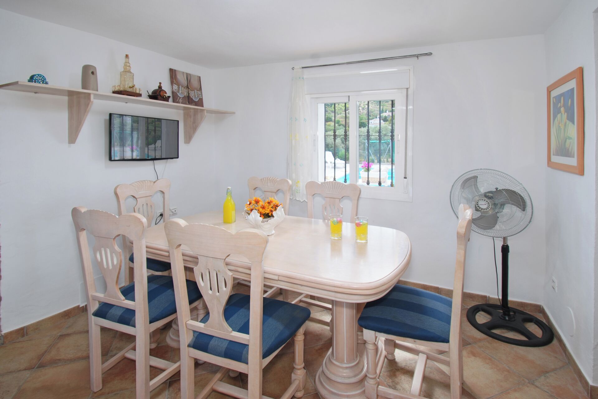 Un comedor con una mesa redonda, seis sillas de madera con cojines azules, un televisor montado en la pared, una ventana con vistas a zonas verdes y un ventilador de pie.