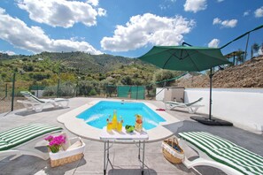 A swimming pool with lounge chairs and umbrellas, surrounded by a fence and hills in the background. - Cosy Poolside Escape for four - Salt-filtered pool. Breathtaking views. (COMARES)