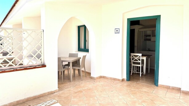 A room with a tiled floor, a table with chairs, a window with a curtain, and a door leading to a kitchen area. - LE RESIDENZE ACQUA DI MARE (Eden Beach)