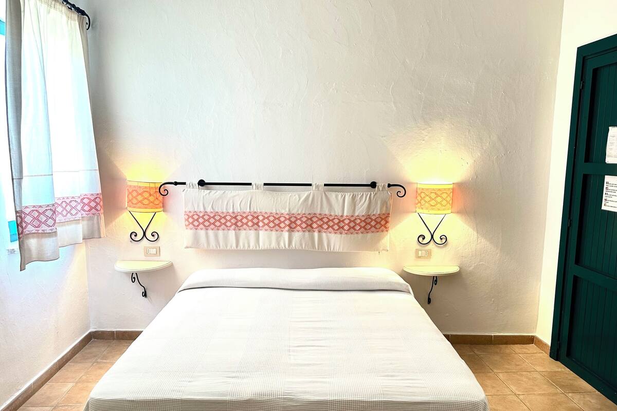 A bedroom with a bed, two bedside lamps, a wall-mounted curtain rod, and a door.