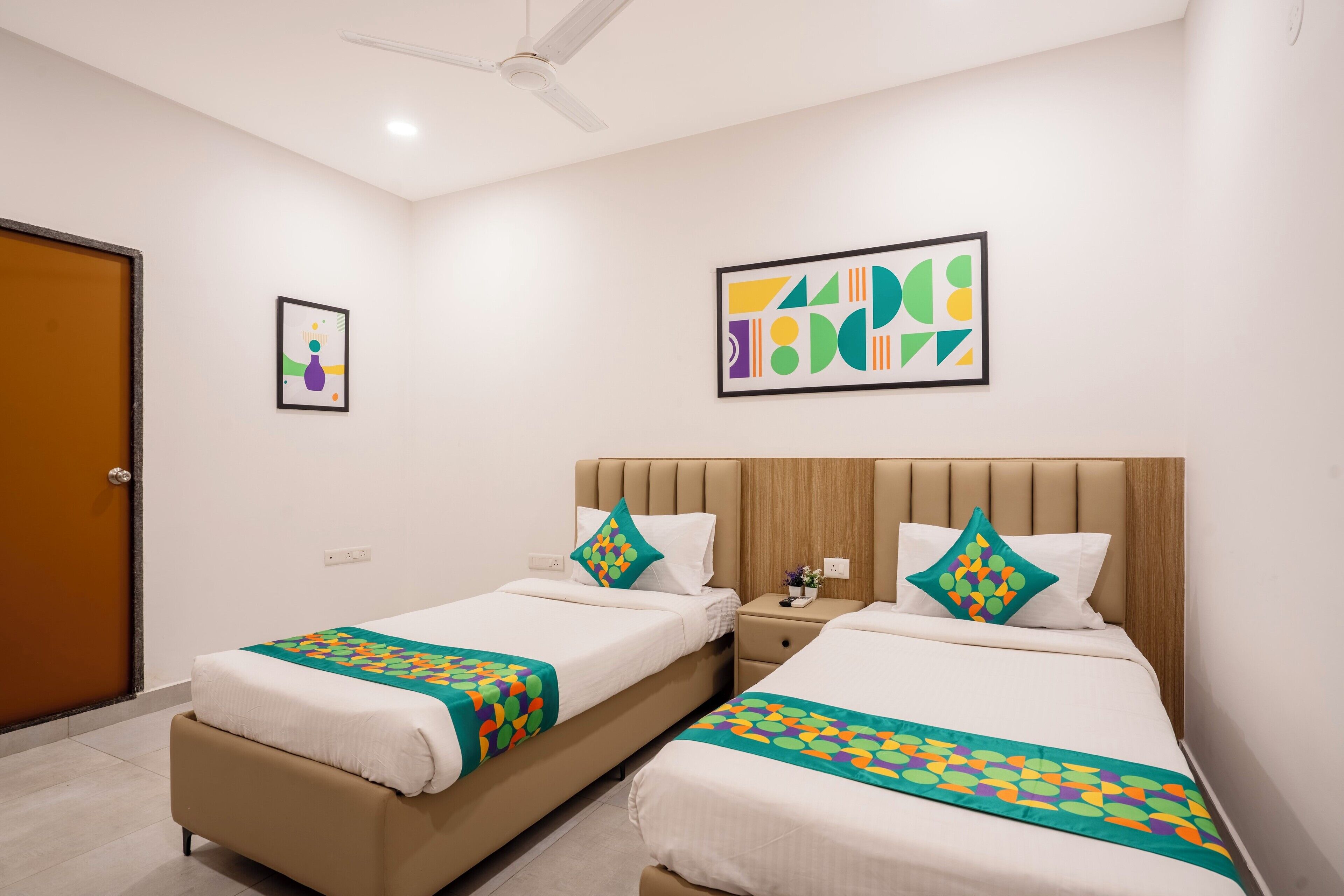 Habitación de hotel con dos camas, cada una con tablero de cabecera color beige y ropa de cama blanca. Hay dos cuadros abstractos enmarcados en las paredes, un ventilador de techo y una puerta con marco marrón.