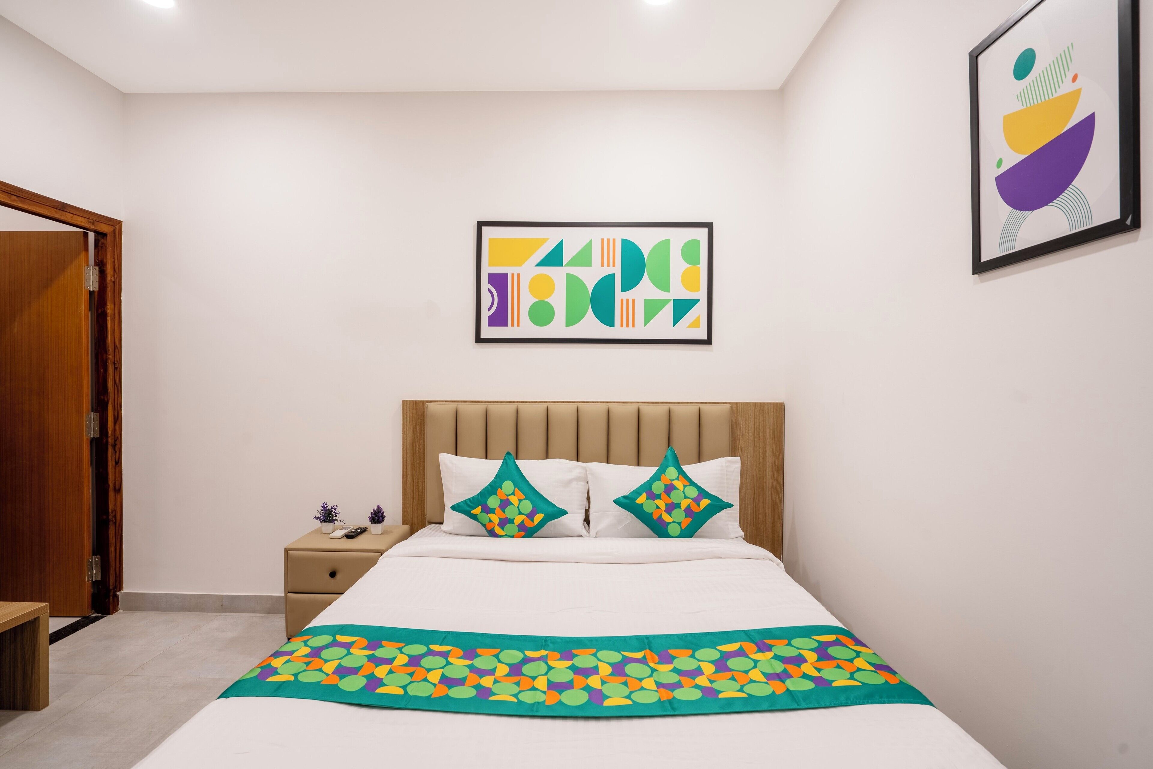 Une chambre à coucher avec un lit, deux oreillers, une table de chevet, un tableau abstrait encadré et une porte.
