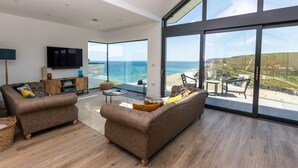 A modern living room with a large window overlooking a beach, a flat-screen TV, and a cozy seating area. - Ocean Skies - sleeps 10 guests  in 4 bedrooms (Porthtowan)