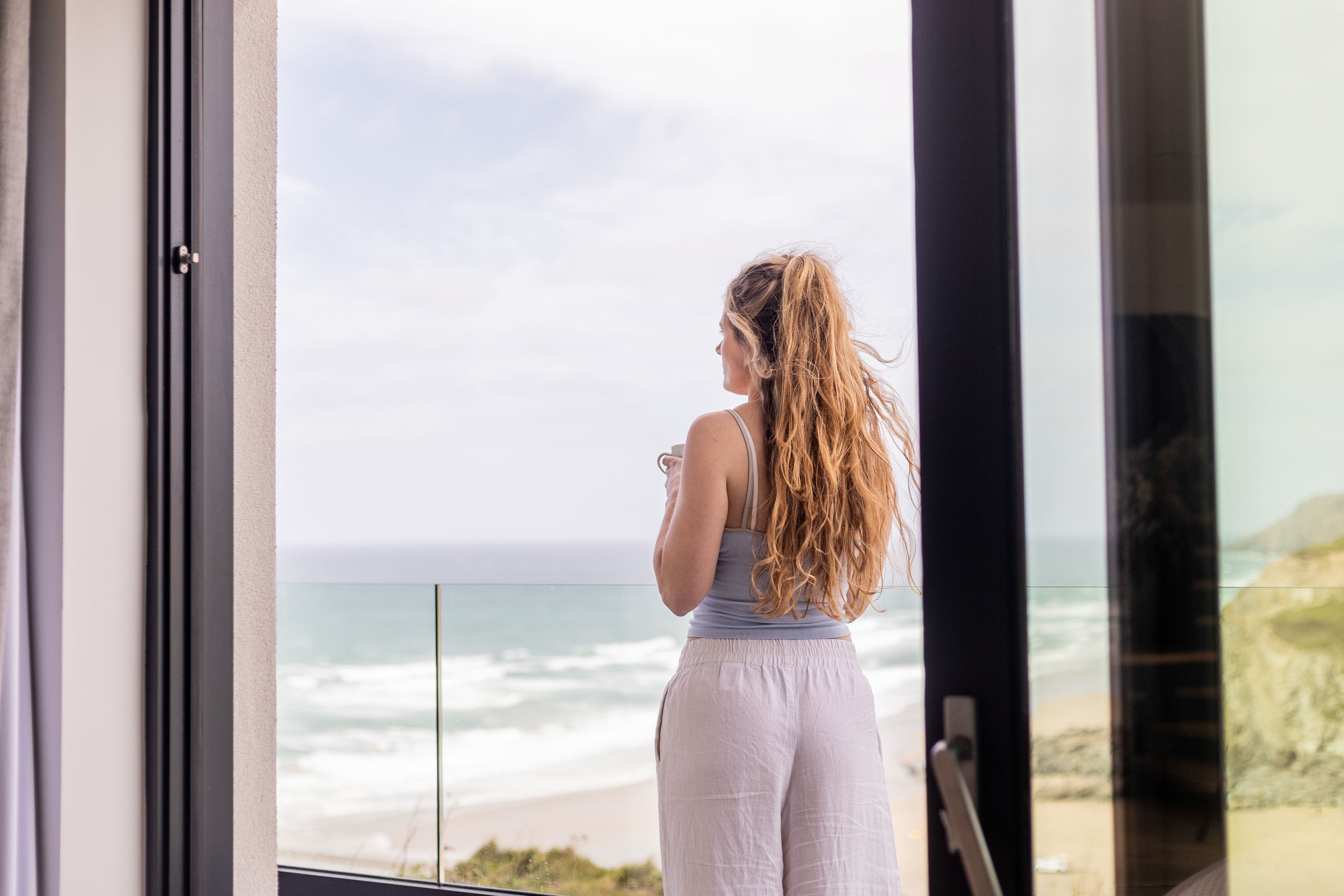 A person standing by a window overlooking a beach.