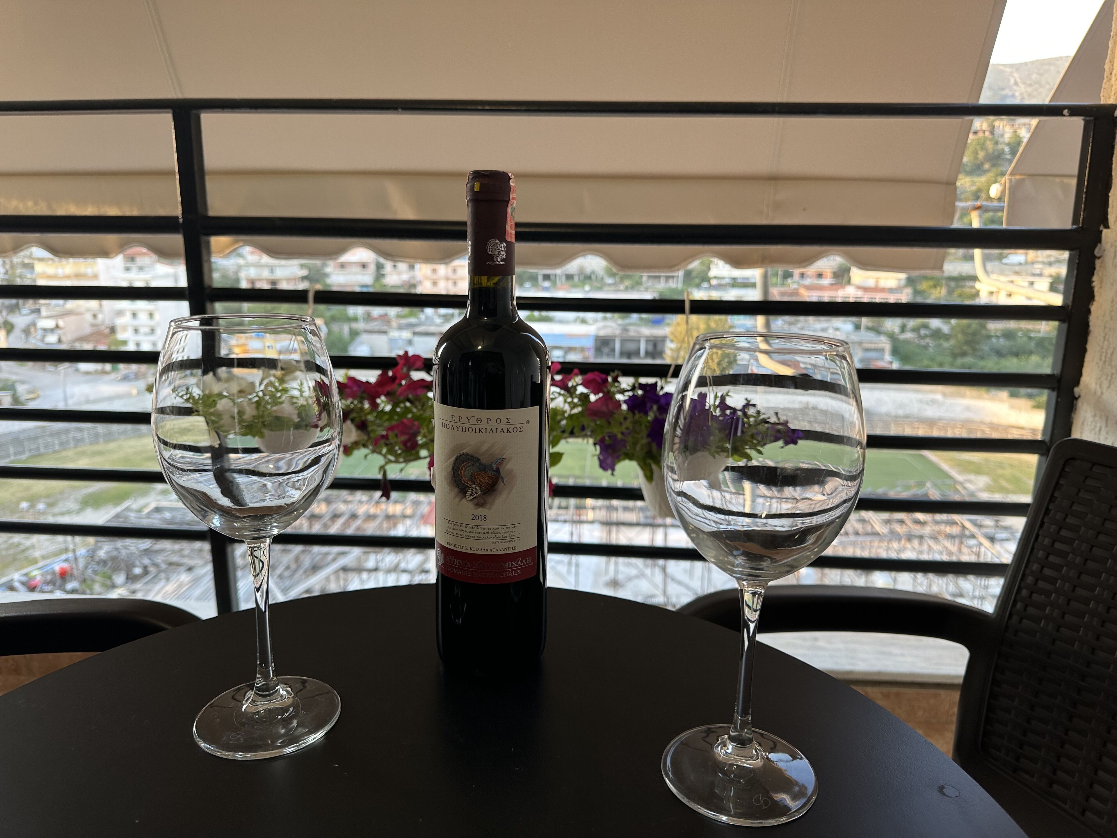 Een tafel met twee wijnglazen en een wijnfles op een balkon, met uitzicht op gebouwen en een tuin.