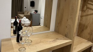Una mesa de madera con botellas de vino y copas, un miniibar gris, y el reflejo de una habitación con un sofá y cortinas.