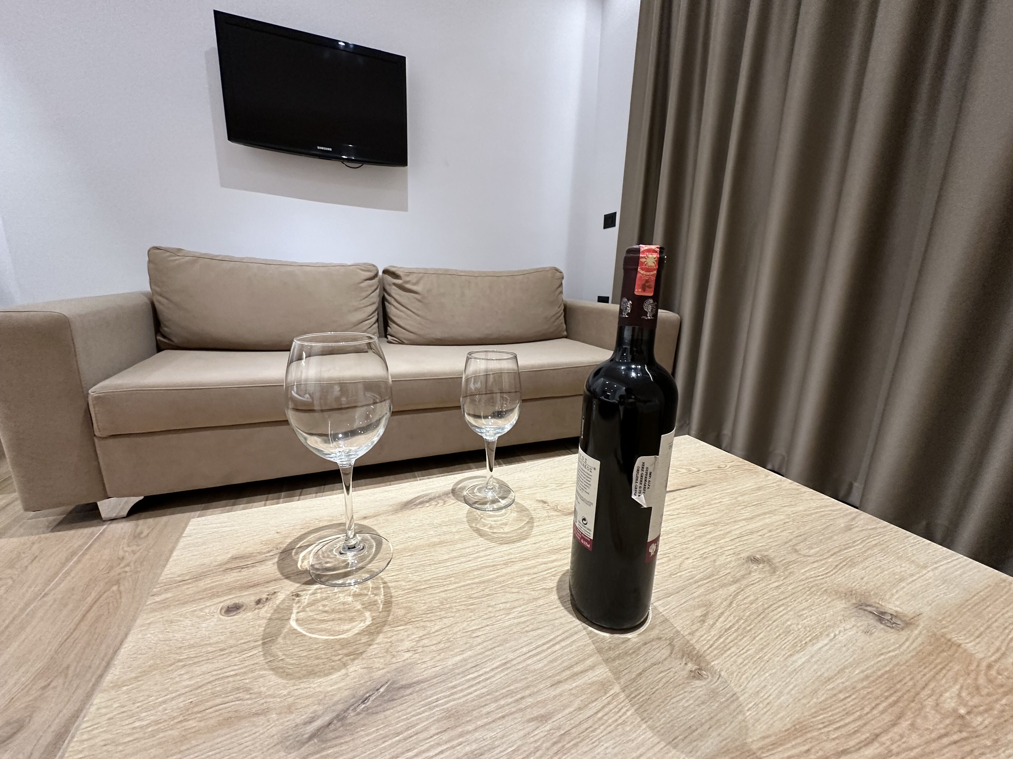 A room with a wooden table, two empty wine glasses, and a bottle of wine. A beige sofa and a flat-screen TV are in the background.